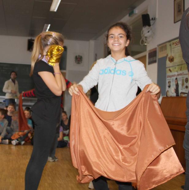 Zwei Mädchen präsentieren ein glänzendes, orangefarbenes Tuch vor einer Gruppe von Kindern.