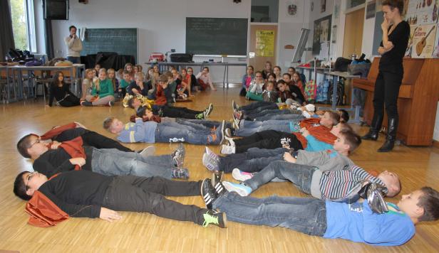 Eine Gruppe Schüler liegt auf dem Boden, während andere zusammensitzen und eine Lehrerin steht.