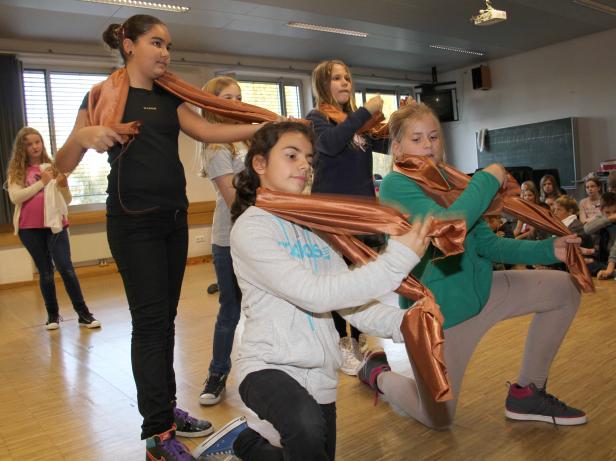 Eine Gruppe von Mädchen führt in einem Klassenzimmer ein Theaterstück mit bronzefarbenen Tüchern auf.