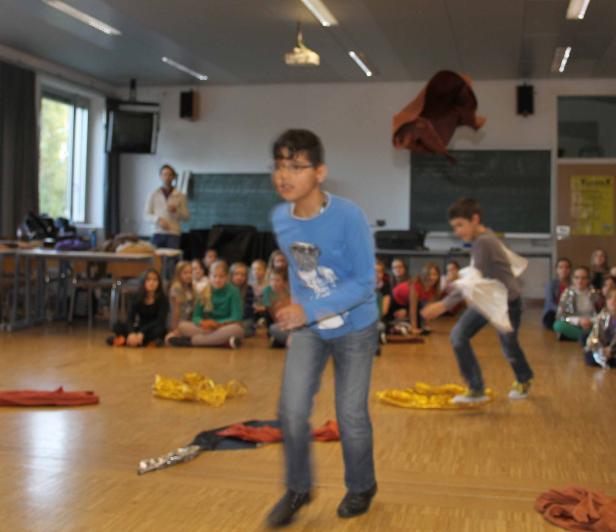 Zwei Jungen führen vor einer Gruppe sitzender Schüler eine Performance mit Tüchern auf.