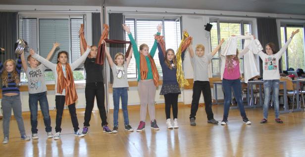 Eine Gruppe von Kindern posiert mit erhobenen Armen und Tüchern in einem Raum.