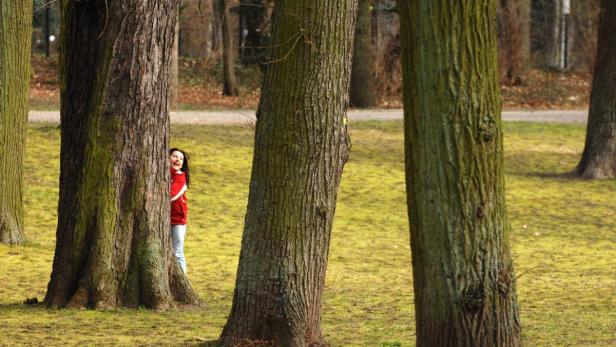 Eine Frau versteckt sich lachend hinter einem Baum im Park.