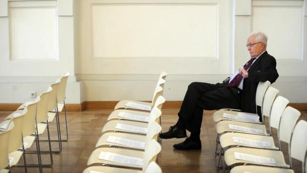 Ein älterer Mann sitzt in einem Anzug in einem leeren Saal auf einem Stuhl.