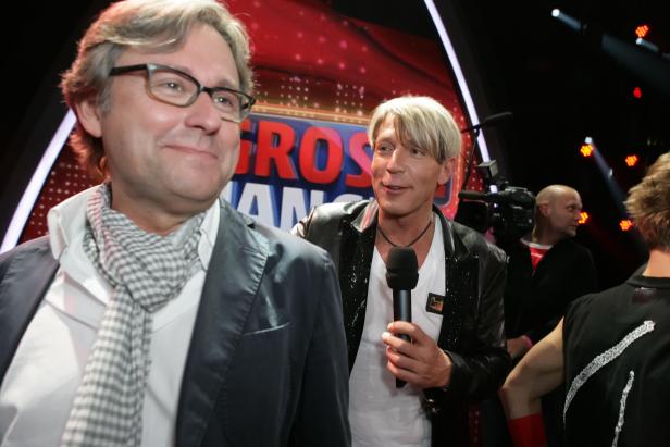 Zwei Männer stehen vor einer Showbühne, einer hält ein Mikrofon.