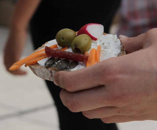 Eine Hand hält ein belegtes Brot mit Oliven, Radieschen, Karotten und Wurst.