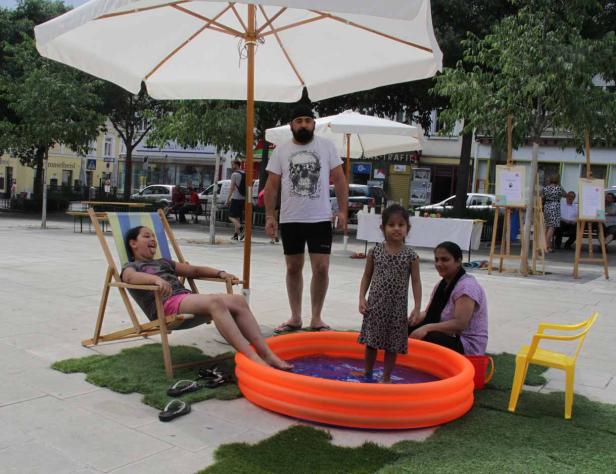 Auf einem Platz planschen Kinder in einem Pool, während ein Mann und eine Frau zusehen.