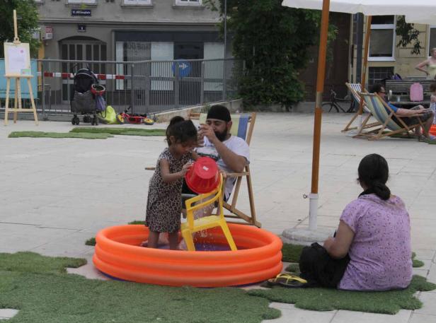 Ein kleines Mädchen spielt mit einem Eimer in einem orangefarbenen Planschbecken, während ein Mann zusieht.