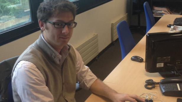 Ein Mann mit Brille sitzt an einem Schreibtisch mit Computer und Telefon.