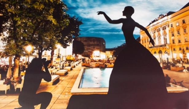Aus der Schattentanz-Show der Wiener Schattenfestspiele von WienTourismus für die Festspielstädte Salzburg und Bregenz (c: wientourismus) Eine Silhouette einer Frau in einem Kleid vor einer belebten Stadtplatzszene.