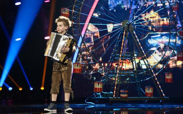 Ein Junge in Lederhosen spielt Akkordeon vor einem beleuchteten Riesenrad.