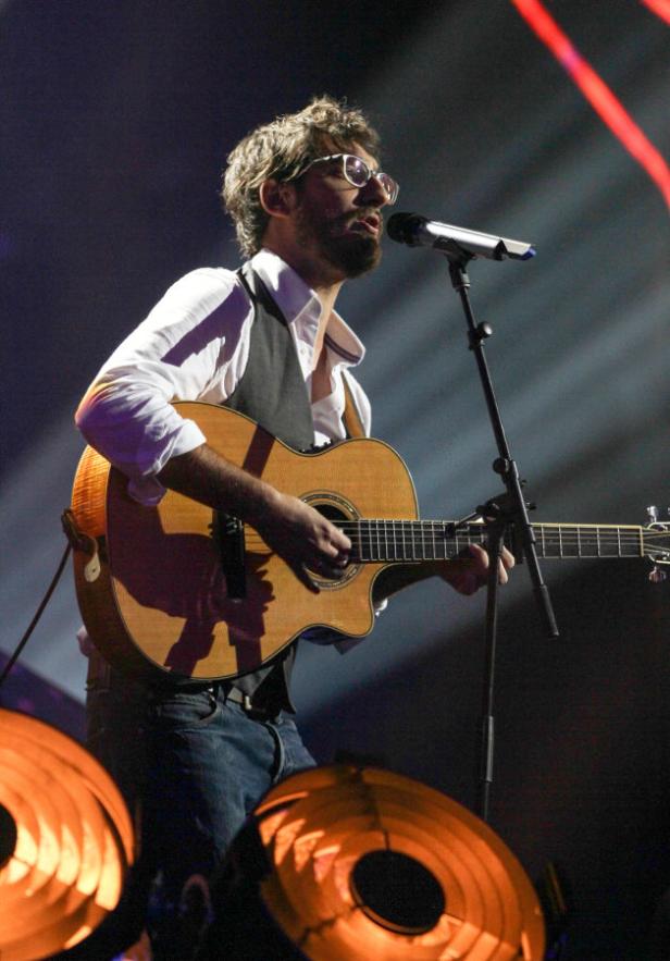 Ein bärtiger Mann mit Brille spielt Gitarre und singt in ein Mikrofon auf einer Bühne.