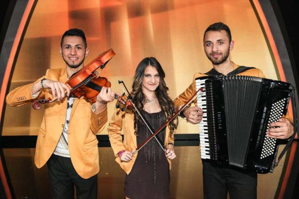 Eine Musikgruppe mit zwei Geigern und einem Akkordeonspieler in goldenen Jacken.