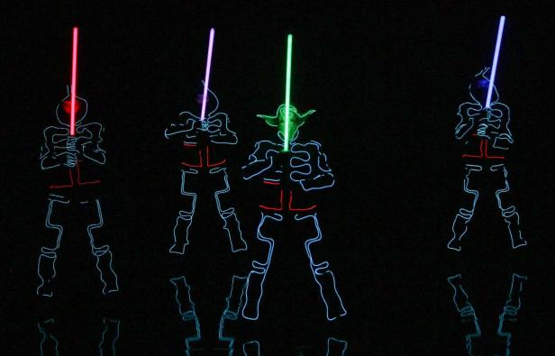Drei Personen in leuchtenden Kostümen, die Lichtschwerter halten, posieren im Dunkeln.