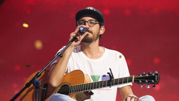 Jason Mraz singt mit Gitarre auf einer Bühne vor rotem Hintergrund.