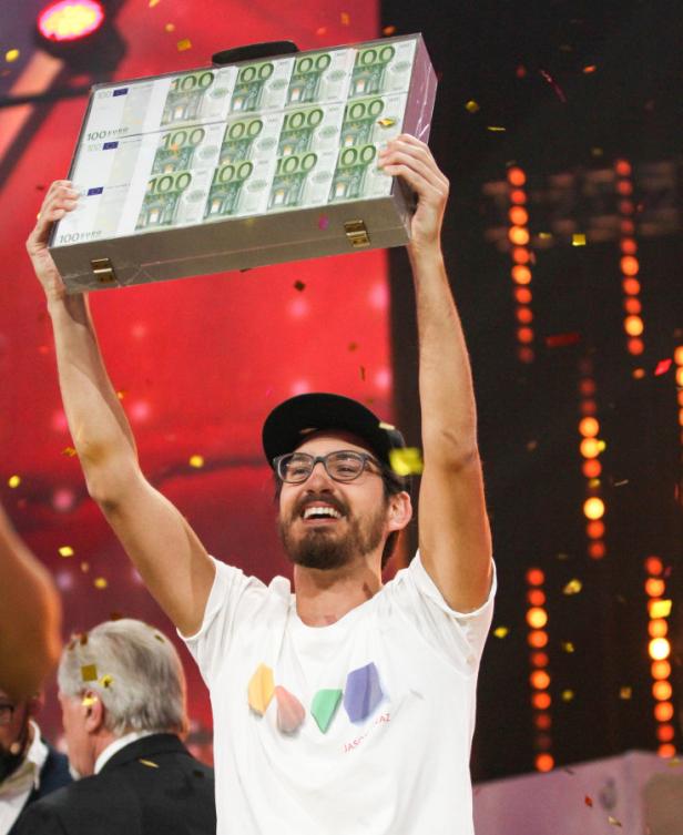 Ein Mann mit Brille und Kappe hält einen Koffer voller 100-Euro-Scheine hoch.