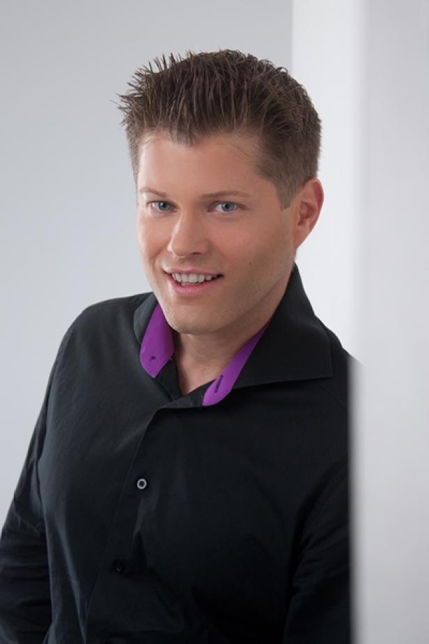 Ein lächelnder Mann mit blonden Haaren und einem schwarzen Hemd mit lila Kragen.