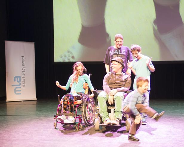 Eine Gruppe von Jugendlichen, von denen zwei im Rollstuhl sitzen, posiert auf einer Bühne für ein Foto.