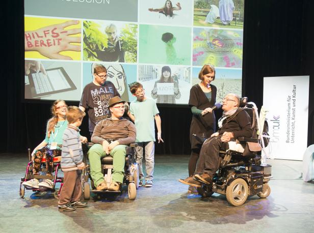 Eine Gruppe von Menschen, darunter einige im Rollstuhl, steht auf einer Bühne vor einer Leinwand.