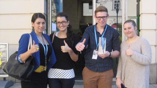 Vier junge Leute stehen vor einem Gebäude und zeigen den Daumen nach oben. Ein Mann hält eine Trophäe.