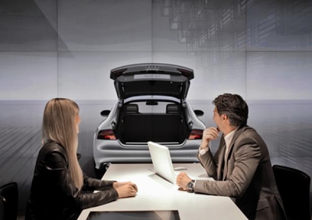 Ein Mann und eine Frau sitzen an einem Tisch vor einem Audi mit geöffnetem Kofferraum.