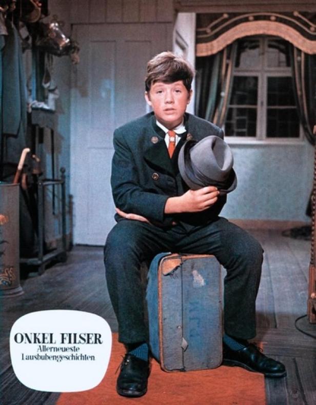 Lausbubengeschichten - Onkel Filser - Hansi Kraus (c: tvspielfilm.de) Ein Junge im Trachtenanzug sitzt auf einem alten Koffer und hält einen Hut.