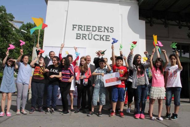 Eine Gruppe von Kindern und Erwachsenen steht vor der Friedensbrücke und hält Papierwindräder hoch.