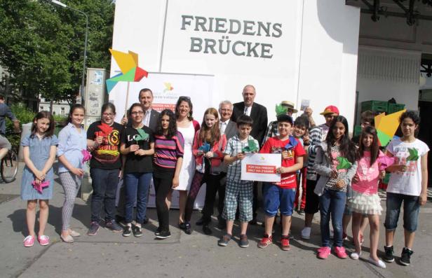 Eine Gruppe von Kindern und Erwachsenen steht vor der Friedensbrücke und hält Papierobjekte und ein Schild.