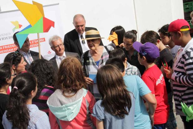Eine ältere Frau mit Hut steht inmitten einer Gruppe von Menschen und hält ein Buch.