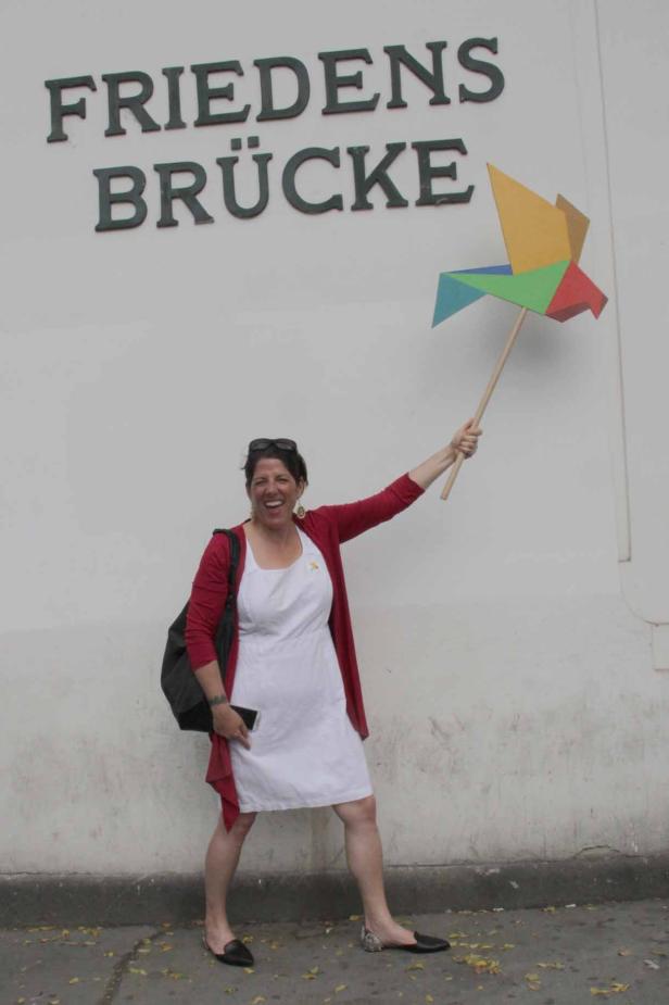 Eine Frau steht vor der Friedensbrücke und hält einen bunten Papierkranich hoch.