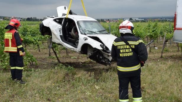 Unfall Weinbergstraße, Porsche… Ein beschädigter weißer Porsche hängt an einem Kran über einem Weinberg, beobachtet von zwei Feuerwehrleuten.