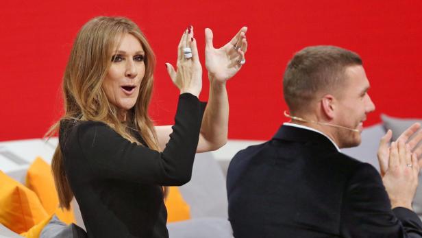 Céline Dion und ein Mann im Anzug klatschen auf einer Couch vor einem roten Hintergrund.