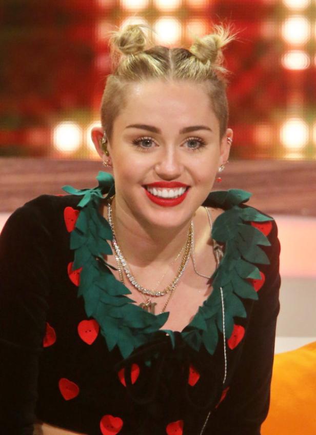 Miley Cyrus lächelt mit roten Lippenstift und Duttfrisur.