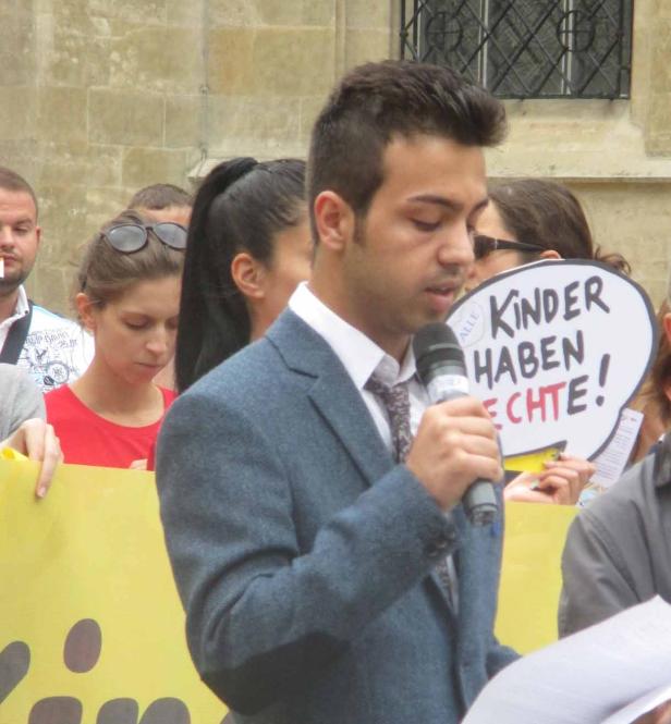 Ein junger Mann spricht bei einer Kundgebung ins Mikrofon. Im Hintergrund ist ein Schild mit der Aufschrift „Kinder haben Rechte!“ zu sehen.