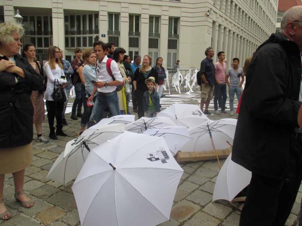 Eine Gruppe von Menschen steht vor einem Gebäude, im Vordergrund liegen weiße Regenschirme.