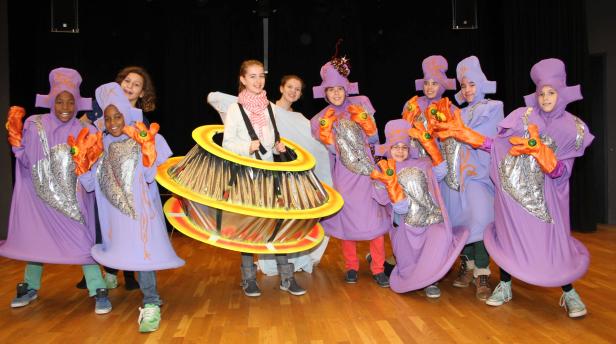 Eine Gruppe Kinder in fantasievollen, lilafarbenen Kostümen steht auf einer Bühne.