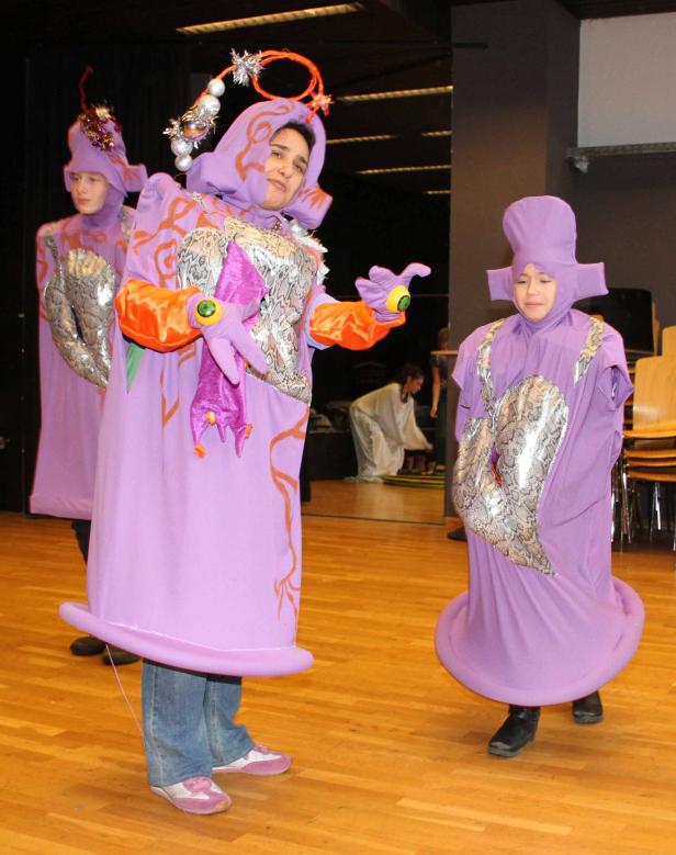 Drei Personen in lila Kostümen stehen auf einer Bühne.