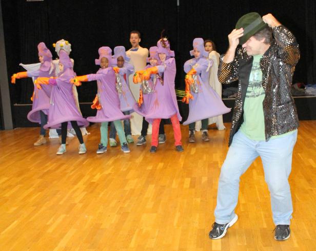Eine Gruppe von Kindern in lila Kostümen tanzt auf einer Bühne, angeleitet von einem Mann mit Hut und Glitzerjacke.