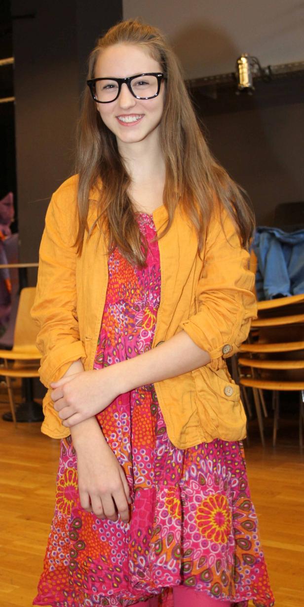 Eine junge Frau mit Brille, gelber Jacke und buntem Kleid lächelt.