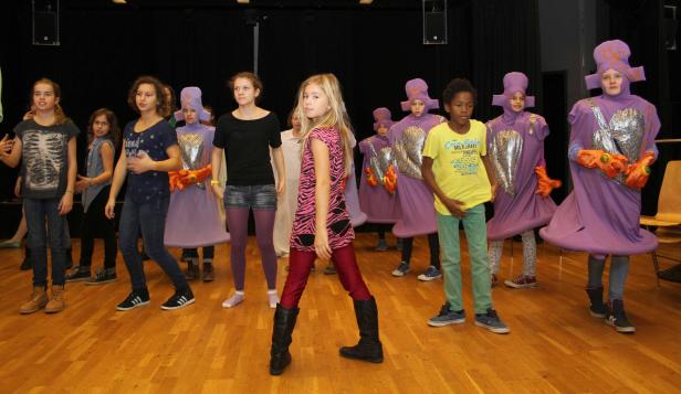 Eine Gruppe von Kindern steht auf einer Bühne, einige in lila Kostümen mit silbernen Brustpanzern.