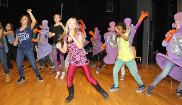 Eine Gruppe von Kindern tanzt auf einer Bühne, einige in lila Kostümen.