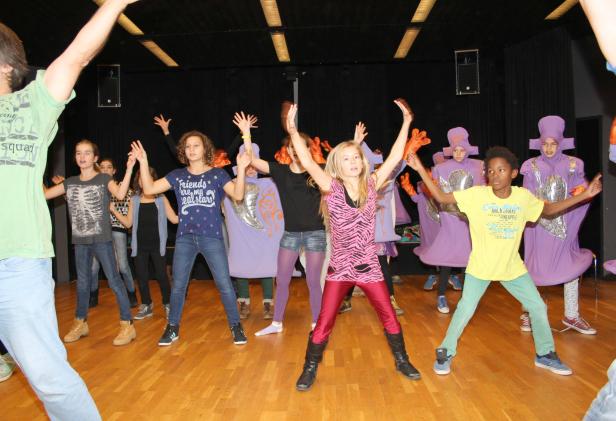 Eine Gruppe von Kindern tanzt auf einer Bühne, einige in lila Kostümen.