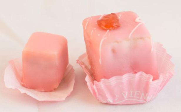 Zwei rosa Petits Fours, einer davon mit kandierter Kirsche, in Papierförmchen.