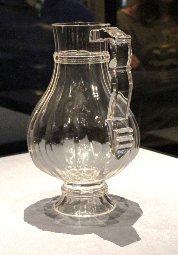 Ein durchsichtiger Glaskrug mit einem verzierten Griff steht auf einem Tisch.