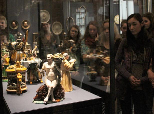 Eine Museumsvitrine zeigt Keramikfiguren und das Spiegelbild von Besuchern.