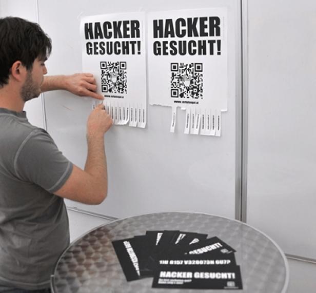 Ein Mann befestigt ein „Hacker gesucht!“-Poster mit QR-Code an einer Wand.