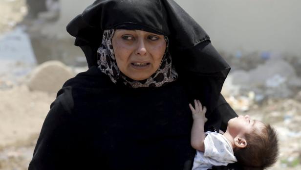 Eine Frau mit Hidschab hält ein Baby vor einem zerstörten Hintergrund.