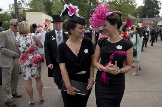 Zwei Frauen mit auffälligen Hüten unterhalten sich bei einem Pferderennen.
