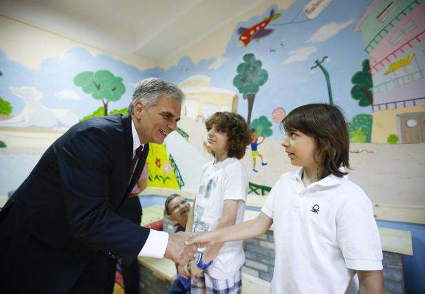 Ein Mann im Anzug schüttelt einem Kind die Hand vor einer Wandmalerei.