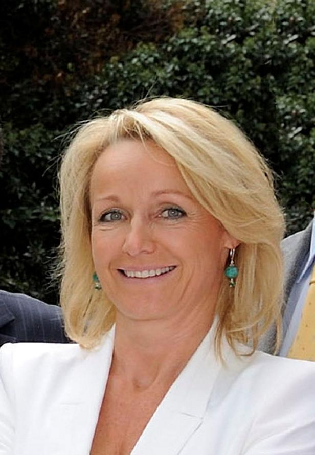Eine lächelnde Frau mit blonden Haaren und einem weißen Blazer.