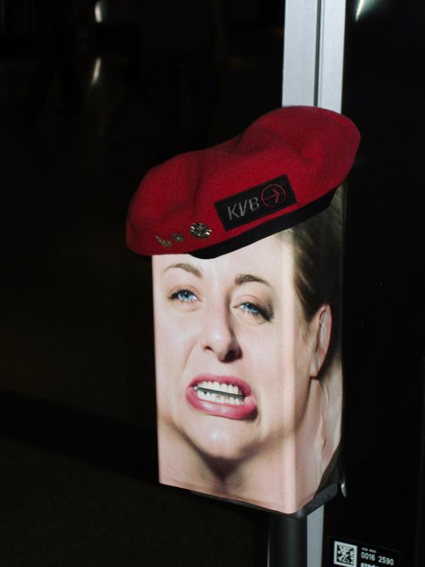 Ein rotes Barett mit dem KVB-Logo sitzt auf einem Plakat mit dem Gesicht einer Frau.